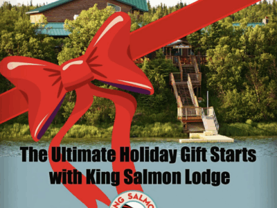 A large red ribbon and bow overlay a photo of King Salmon Lodge on the Naknek River, with stairs leading down to the water and trees surrounding the lodge. Text reads, “The Ultimate Holiday Gift Starts with King Salmon Lodge,” along with the King Salmon Lodge logo.